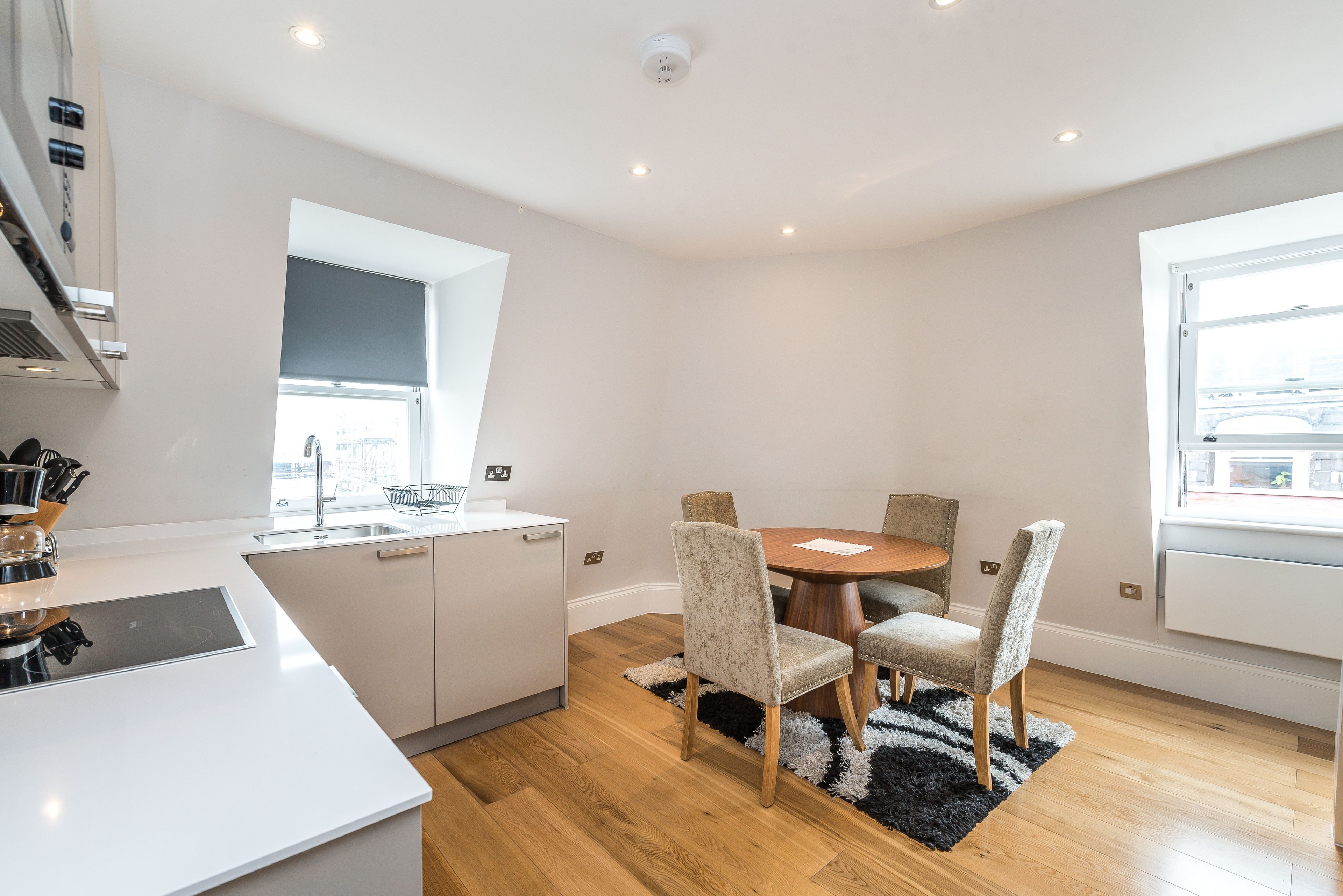 A modern kitchen with white cabinetry, a built-in oven, and a window above the sink. There is a dining area with a round table and four chairs, and a black and white rug on the wooden floor.