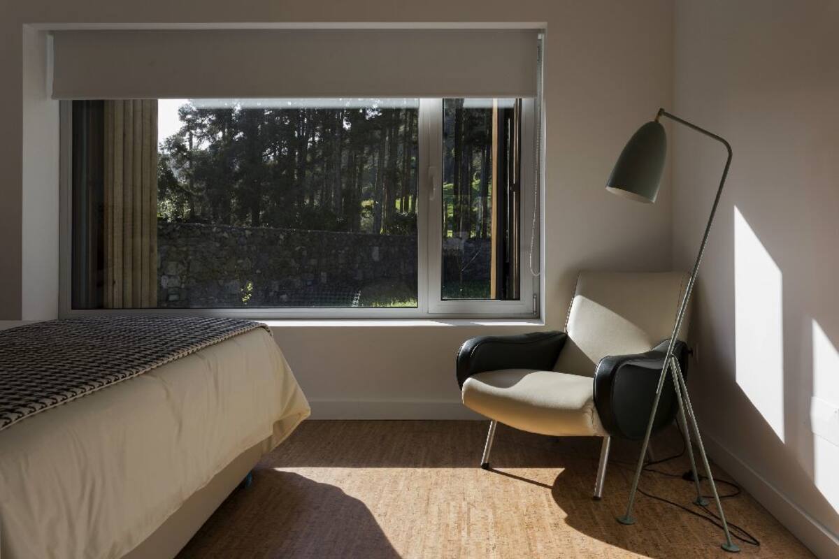 A bedroom with a bed, a chair, a floor lamp, and a window with a view of trees and a stone wall.