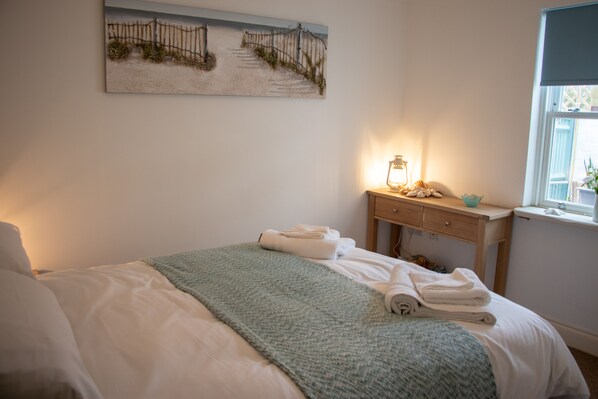 A neatly made bed with a green blanket, a bedside table with a lamp, and a painting of a beach scene on the wall.