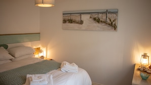 Una cama bien hecha con ropa de cama blanca y una toalla doblada, un cuadro de una playa en la pared, y dos lámparas de pared a cada lado de la cama.