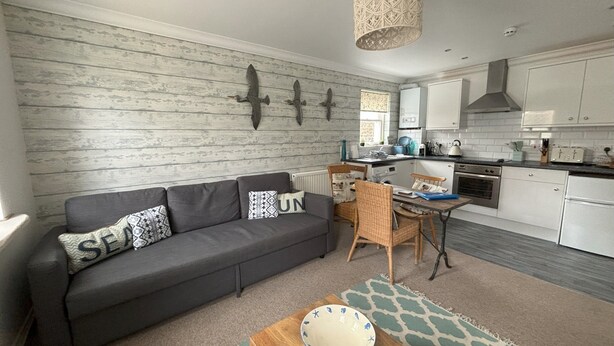 Una cocina moderna con armarios blancos, una zona de comedor con mesa y sillas de madera, y un sofá gris con almohadones decorativos.