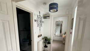 Un pasillo con suelo de madera, paredes blancas, un adorno de brújula en la pared, una lámpara de techo azul y un espejo con marco decorativo.