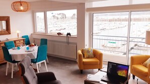 Un coin repas avec une table dressée pour deux personnes, un fauteuil jaune et une vue sur une marina, visible à travers de grandes fenêtres.