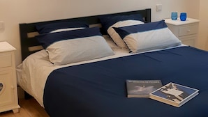 Un lit bien fait, recouvert d’une couvre-lit bleu foncé et agrémenté d’oreillers, une table de chevet avec deux verres bleus, et un tableau représentant une scène de plage, accroché au mur.