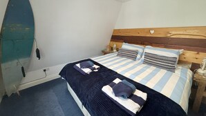 A bedroom with a bed, a wooden headboard, a surfboard leaning against the wall, and a nightstand with a lamp. - Pebbles. The Beach House, apartment in central Eastbourne with private parking (Eastbourne)