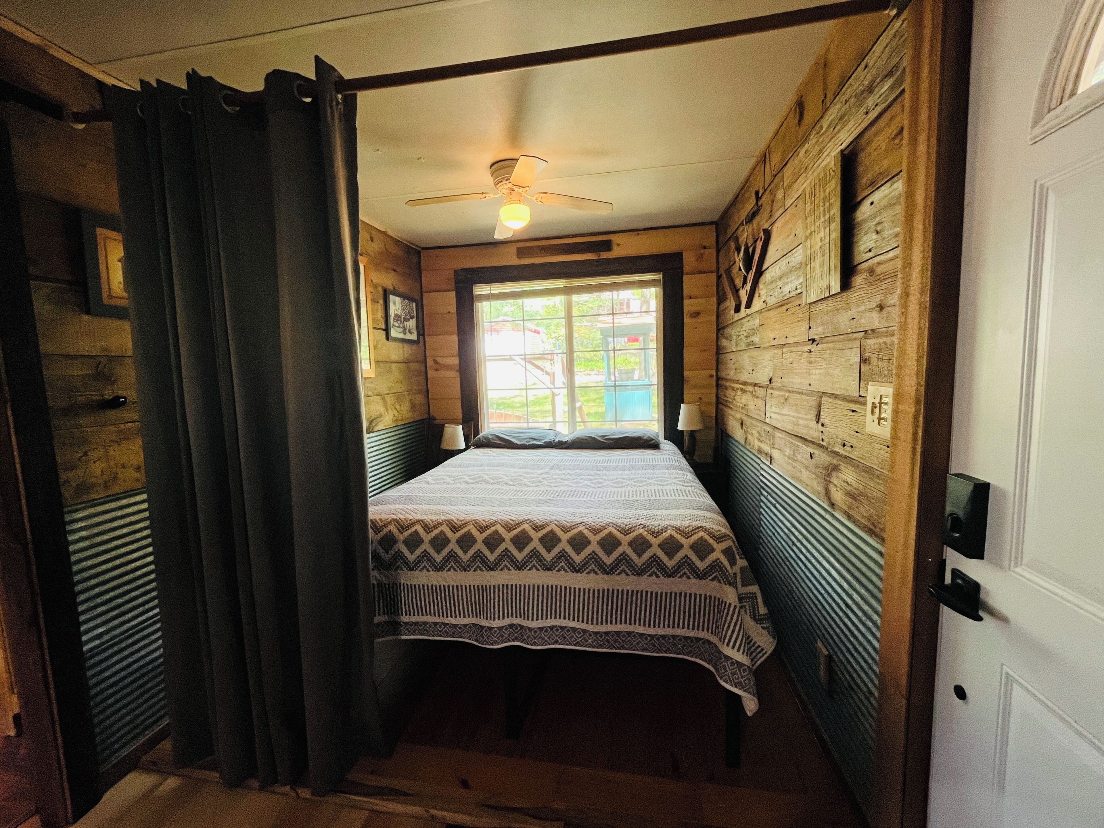 A narrow room with a bed, a ceiling fan, and wooden walls.