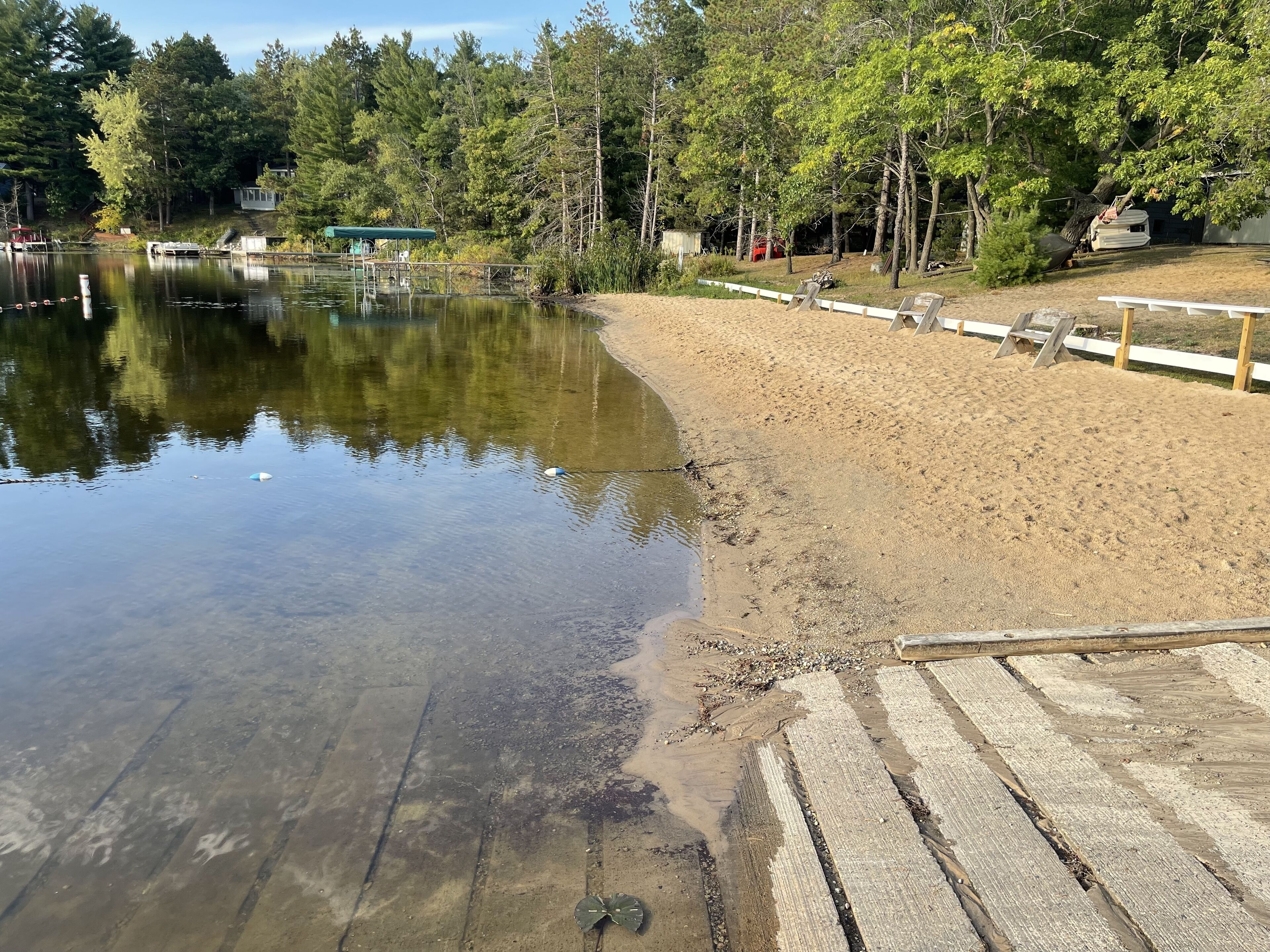 Une plage de sable avec une jetée en bois qui s’avance sur un lac paisible, le tout entouré d’une forêt dense.