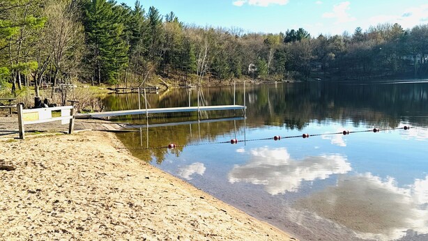 A sandy beach with a wooden bench, a metal dock extending into a calm lake, and a forested shoreline. - WI Dells- full Rec lake,swim,boat,ski,UTV,fish,BBQ,sauna,fire pit,hiking,kayak (Oxford)