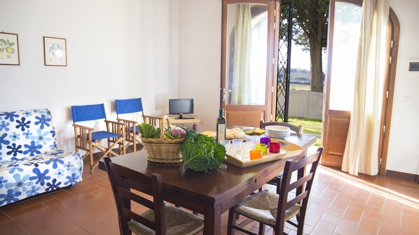 A dining area with a table set for four, a wicker basket with vegetables, a television, and a view of the outdoors.