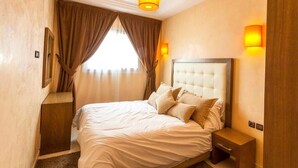 Een hotelkamer met een groot bed, een houten nachtkastje, een wandlamp en een raam met gordijnen.