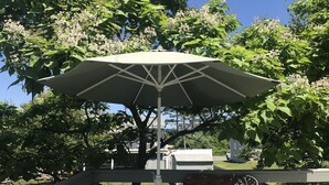 Een terras buiten met een ronde tafel en stoelen, een witte parasol en een houten omheining.