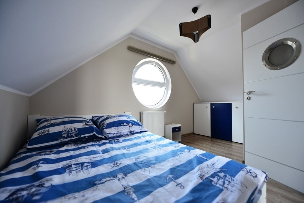 A bedroom with a sloped ceiling, a bed with a blue and white striped bedspread, a round window, a white radiator, a small white cabinet, and a wardrobe with a circular mirror.