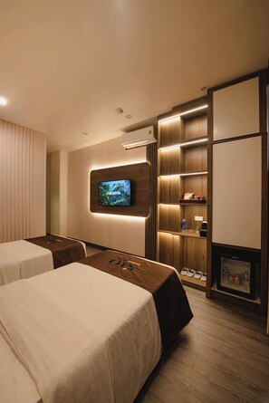 Habitación de hotel con dos camas, un televisor montado en la pared, un estante con refrigerios y un zapatero.