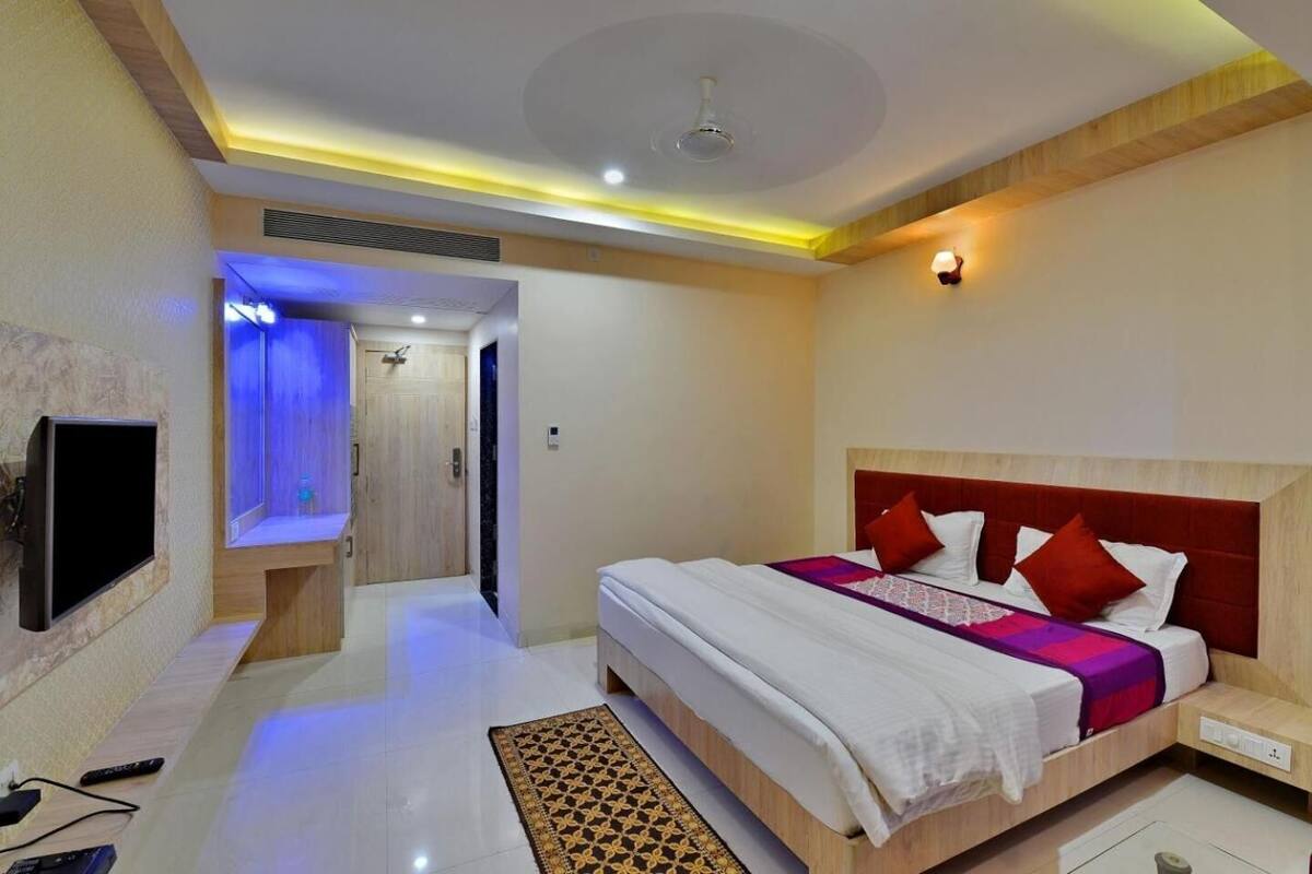 Quarto de hotel com cama, televisão, banheiro com chuveiro e corredor.