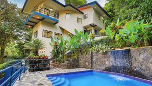 Un moderno edificio de dos pisos con piscina, rodeado de exuberante vegetación y una cascada de piedra.