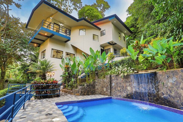 A modern two-story building with a swimming pool, surrounded by lush greenery and a stone waterfall feature.