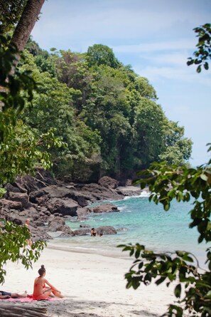 Ein Strand mit einer Person, die auf einer Handtuch liegt, umgeben von üppiger Vegetation und einer felsigen Küstenlinie.