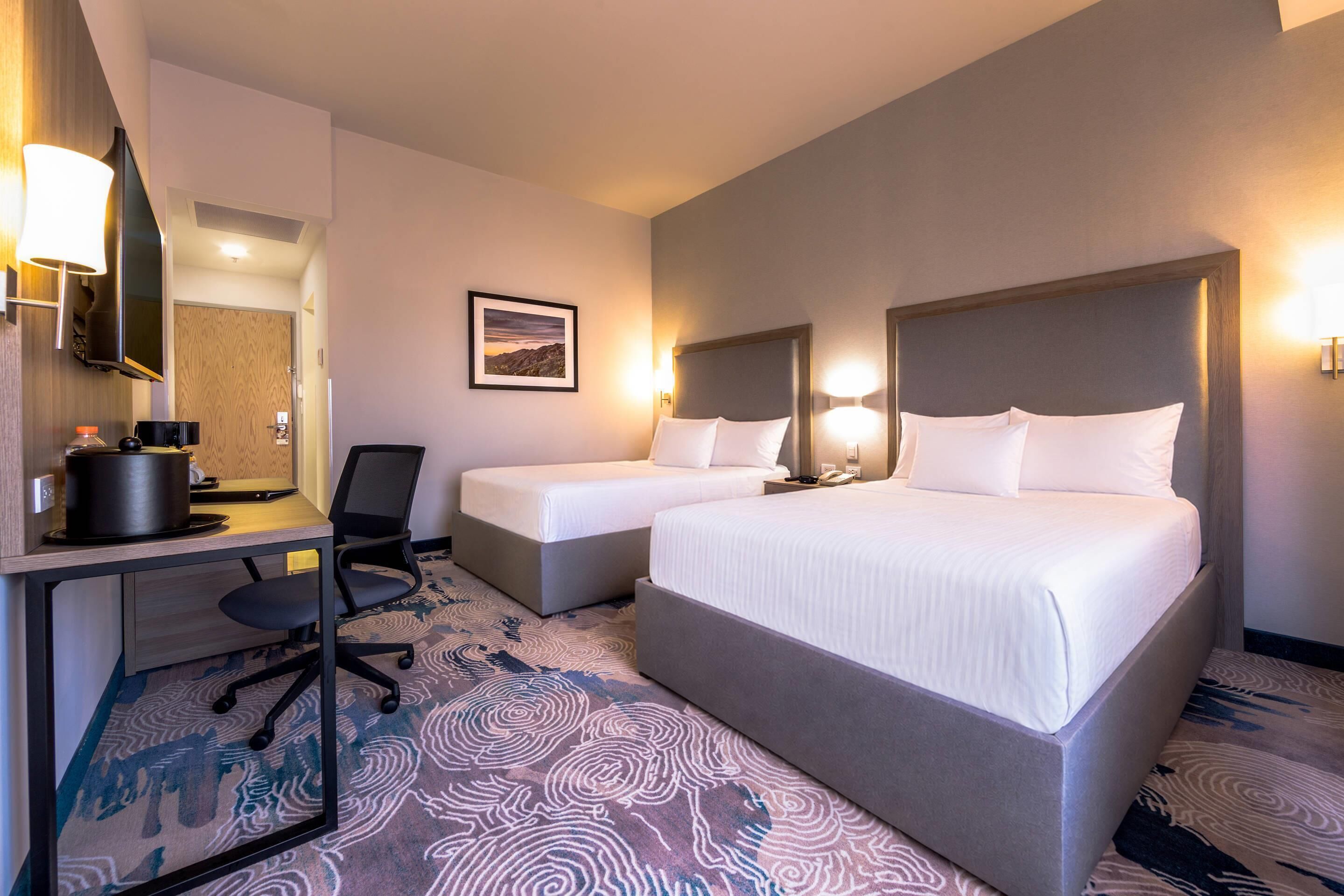 A hotel room with two beds, a desk with a chair, a flat-screen TV, and a framed picture on the wall.