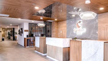 Zona de recepción de un hotel con un mostrador de madera, un centro de negocios y un cartel que dice 'Comfort Inn'.