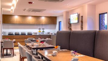 A modern dining area with tables and chairs, a kitchen in the background, and a television on the wall.