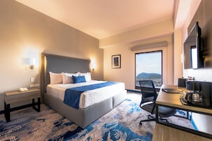 Habitación de hotel con cama, mesita de noche, escritorio, silla y un amplio ventanal con vistas.