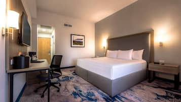 A hotel room with a bed, desk, chair, and a small bedside table with a lamp.