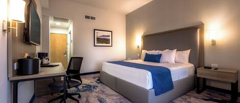 A hotel room with a bed, desk, chair, and a small table with a lamp.