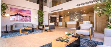 A modern hotel lobby with a high ceiling, a reception desk, seating area, and a large painting.