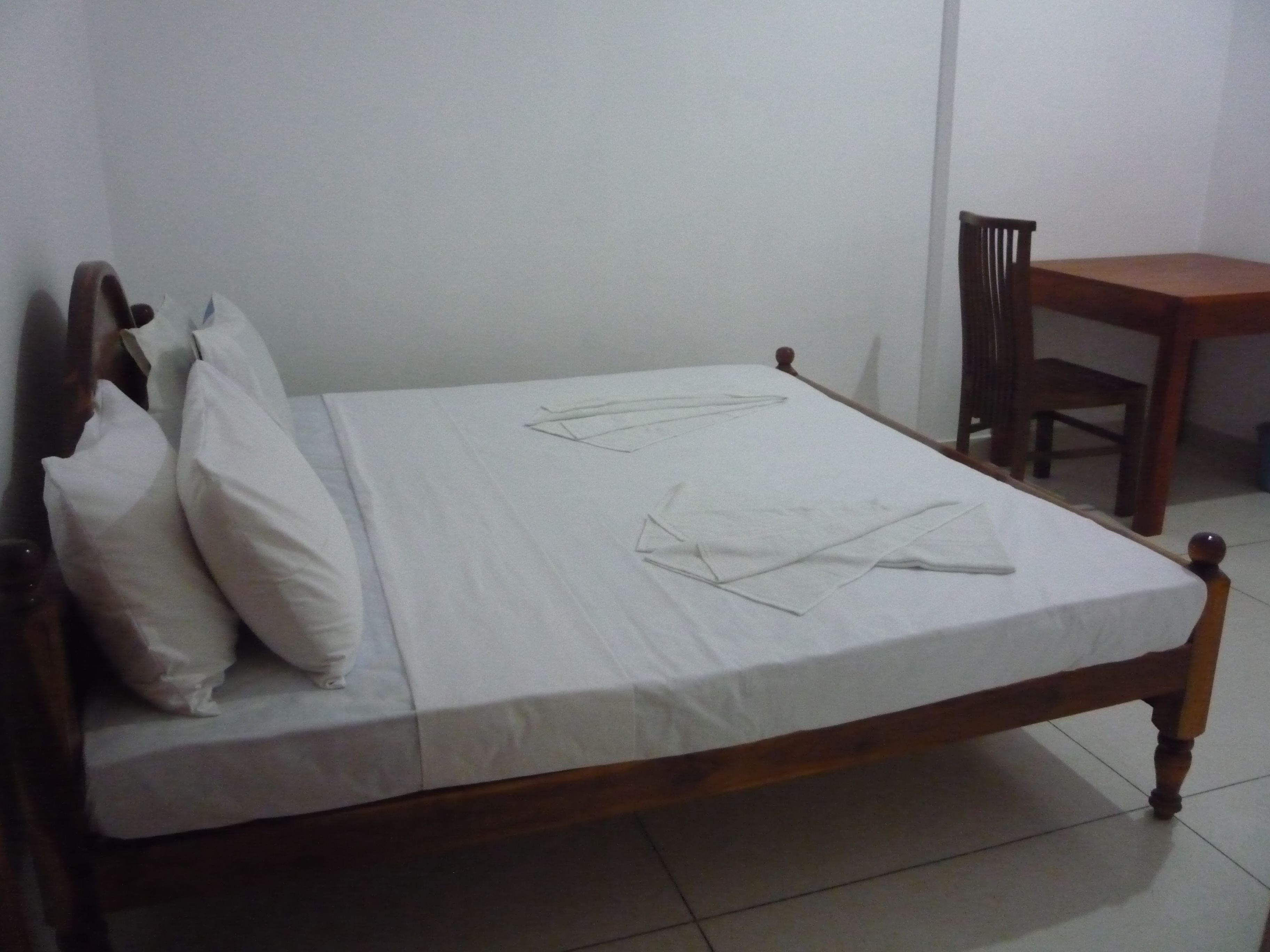 A single bed with a wooden frame and white bedding, a wooden chair, and a small table in a room with plain walls and tiled floor.