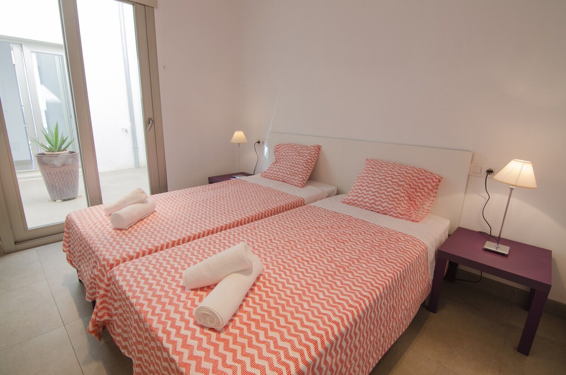 A hotel room with two beds, each with a patterned bedspread, pillows, and rolled towels. There are bedside tables with lamps and a potted plant visible through the glass door.