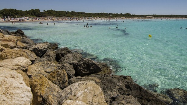 Una playa con agua turquesa cristalina, costa rocosa y una zona de playa concurrida al fondo.