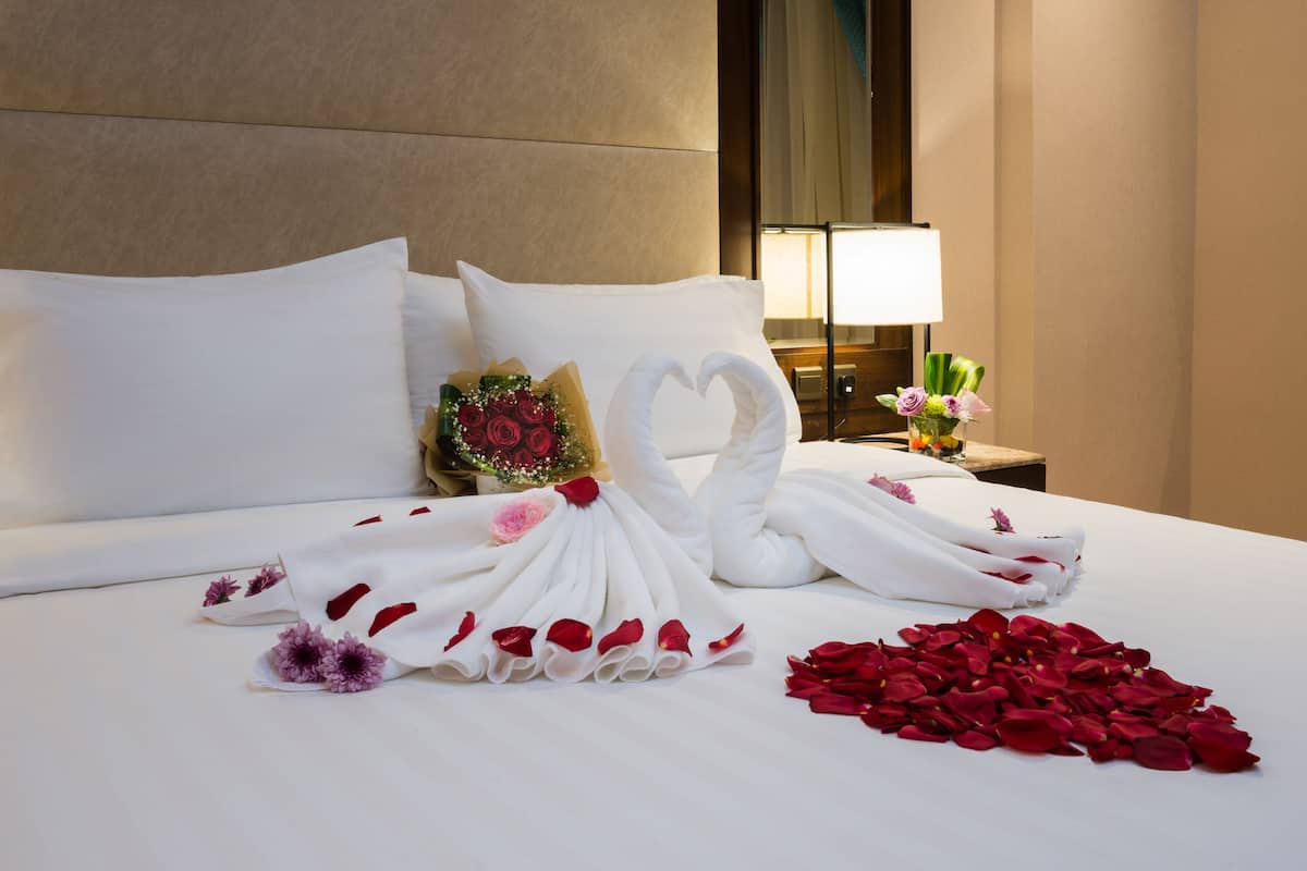 A hotel room with a neatly made bed, a bouquet of flowers, and a heart-shaped towel arrangement.
