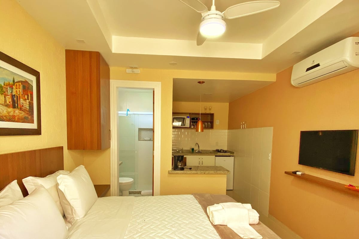 A hotel room with a bed, a ceiling fan, an air conditioning unit, a television, and a small kitchenette.