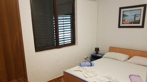 A bedroom with a wooden bed, a nightstand with a lamp, a window with blinds, and a framed picture on the wall. - Economy double room - close to the Velika Plaza for cheap price (UL)