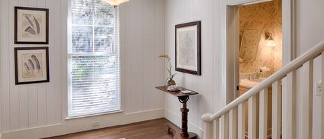 A hallway with wooden flooring, a white railing, and a staircase leading to a bathroom with a floral wallpaper design.