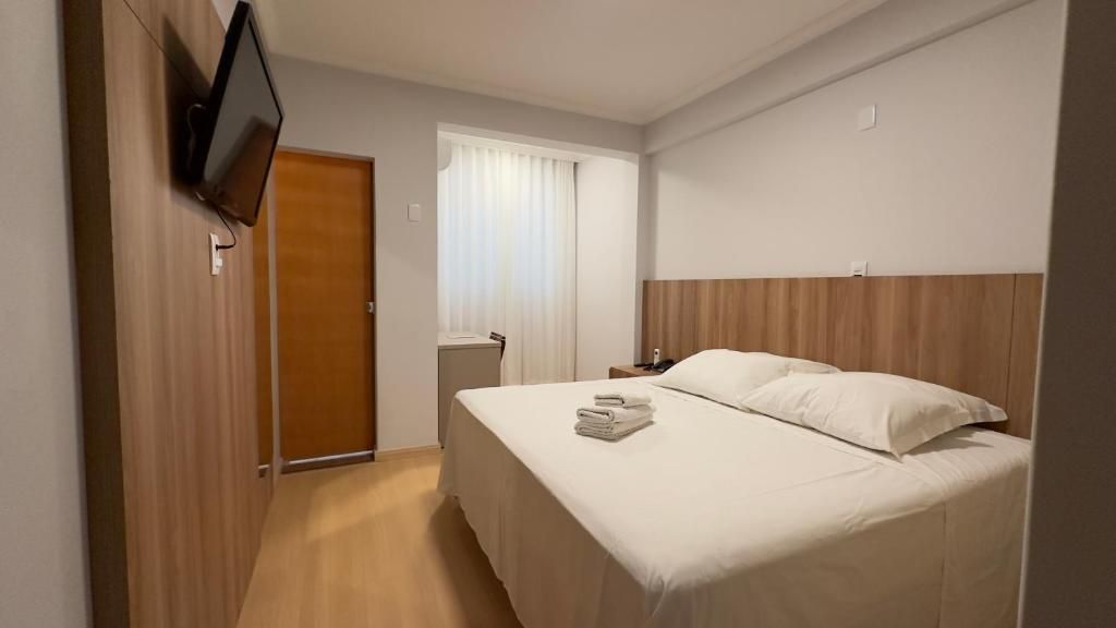 Quarto de hotel com cama, televisão fixada na parede, janela com cortinas e porta de madeira.