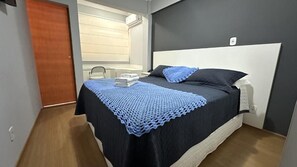 A bedroom with a bed, a bedside table, a window with blinds, and a door.
