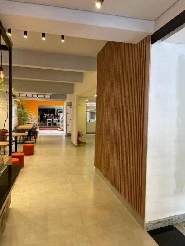 Un pasillo con pared de paneles de madera, suelo de baldosas y una zona de estar con mesas y sillas al fondo.