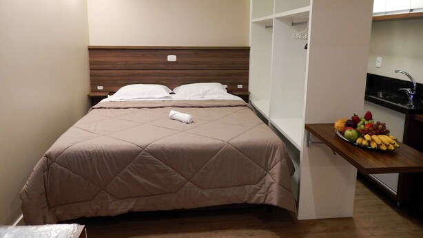 A compact hotel room with a bed, bedside table, and a small kitchenette area with a fruit bowl. - Flats Requinte Gramado (Gramado)