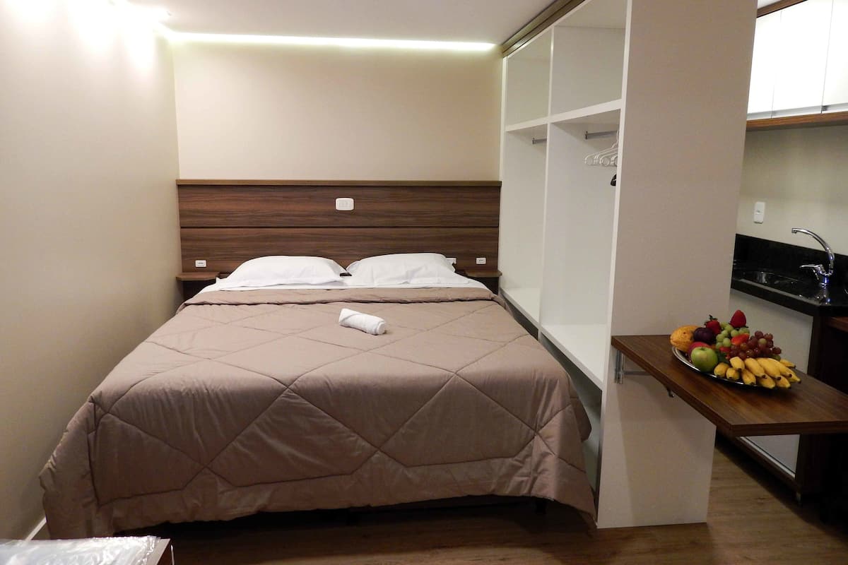 Quarto de hotel compacto com cama, mesa de cabeceira e pequena área de cozinha equipada, com uma fruteira.