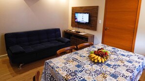 A living room with a dark blue sofa, a wooden dining table with a patterned tablecloth, and a fruit bowl on the table. - Flats Requinte Gramado (Gramado)