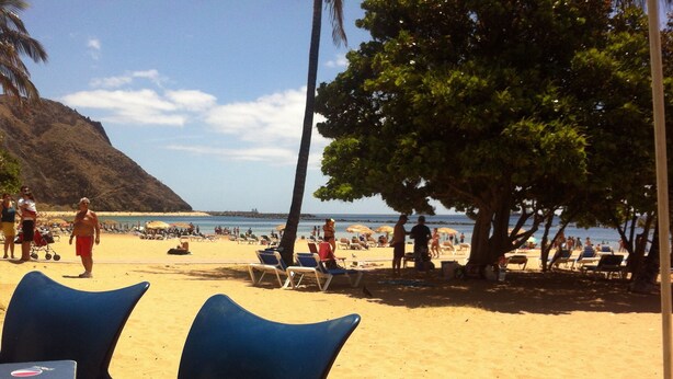 A beach with blue chairs and a table, a palm tree, and people in the background. - Casa "MANUELA" Feel at home in La Paz - Vivienda Vacacional (Puerto de la Cruz)