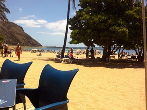 Een strand met blauwe stoelen en een tafel, een palmboom en mensen op de achtergrond.