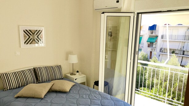 Un dormitorio con una cama, una mesita de noche con lámpara, un cuadro en la pared y un ventanal corredero que da a un balcón con vistas a edificios.