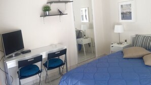Un dormitorio con cama, un escritorio con computadora, una silla, una mesita de noche con lámpara y un estante con planta y figura de dinosaurio.