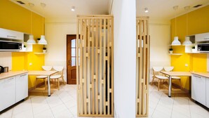 A modern kitchen with yellow walls, white cabinets, and wooden partitions. There are two white dining tables with chairs, and pendant lights above.