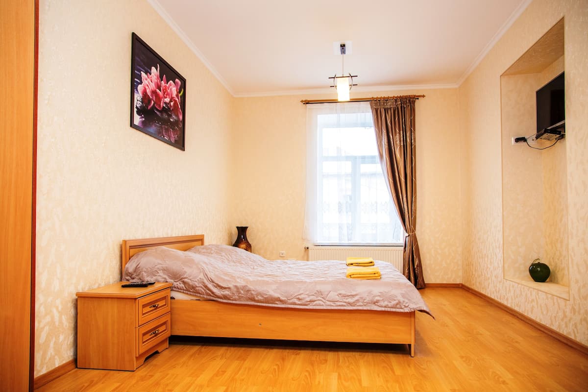 Un dormitorio con una cama de madera, una mesita de noche, un florero, una ventana con cortinas y un televisor montado en la pared.