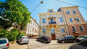 A street view with parked cars, a building with a balcony, and a tree.