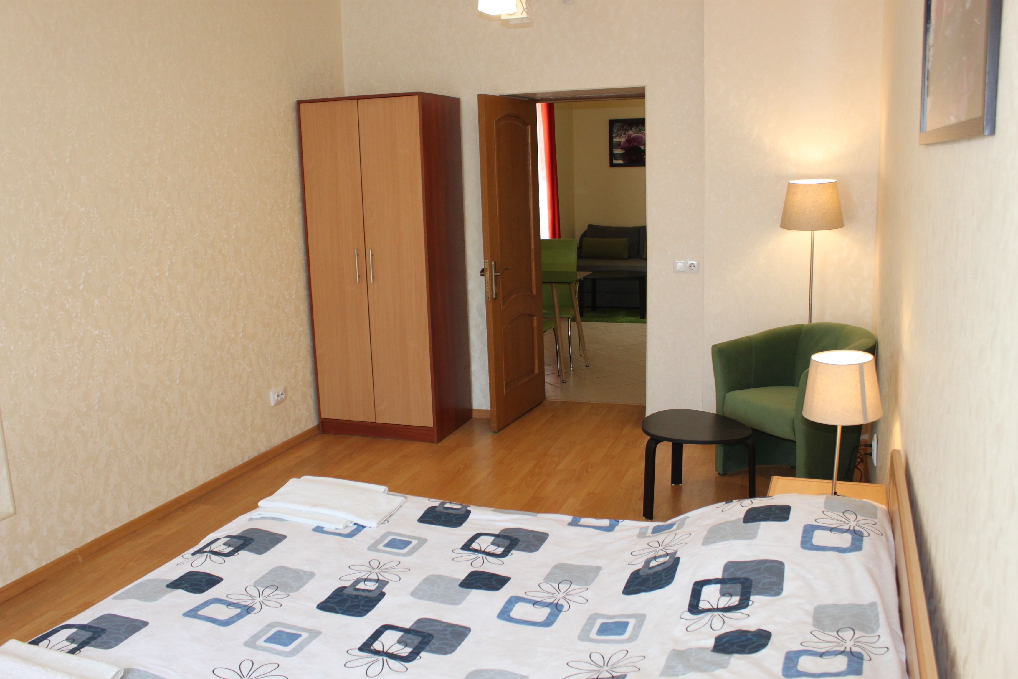 A hotel room with a bed, a wardrobe, a green armchair, a small table, and a lamp.