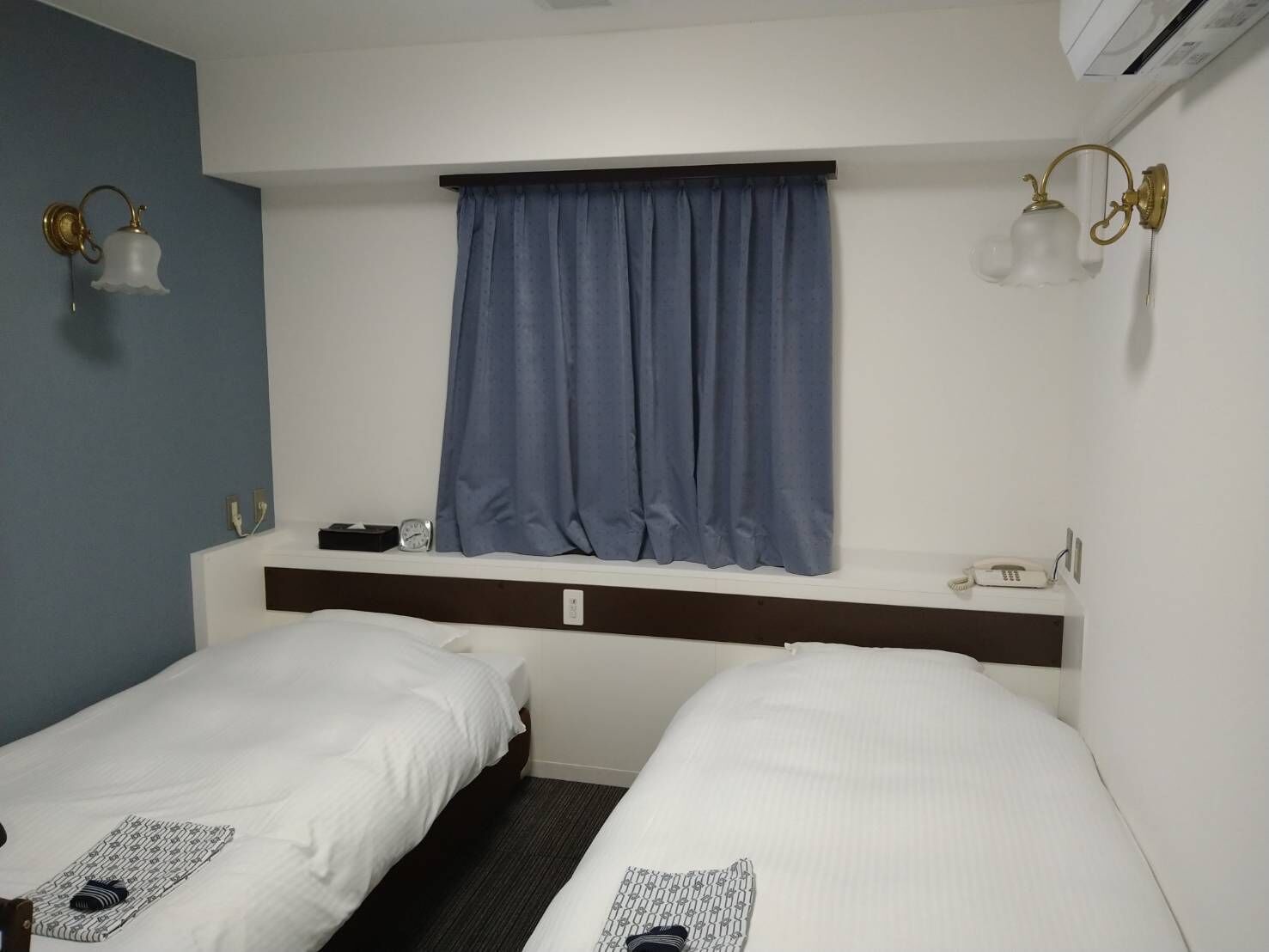 A hotel room with two single beds, a window with a blue curtain, a bedside table with a phone, and wall-mounted light fixtures.